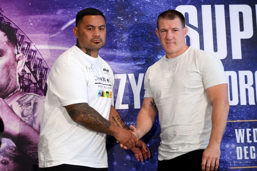 Mark Hunt and Paul Galen stand and shake hands, both wearing white T-shirts against a blue background.