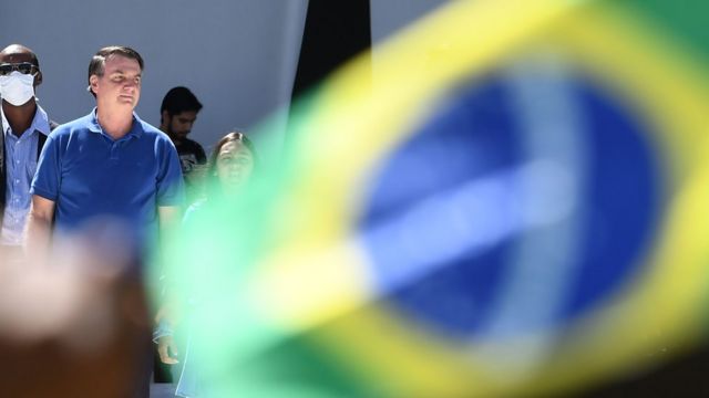 Bolsonaro is seen behind the Brazilian flag and with masked security