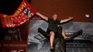 A Liverpool fan celebrates winning the Premier League as he sits on a statue of Bill Shankly outside Anfield