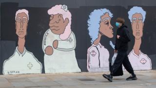 A man wearing a mask walks past a mural of BAME medical and transport workers