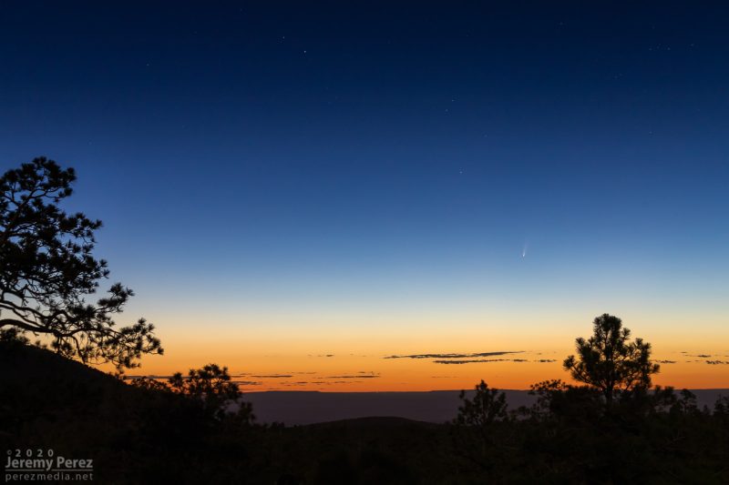 A small faint comet in a twilight sky, with a desert landscape silhouetted in the foreground.