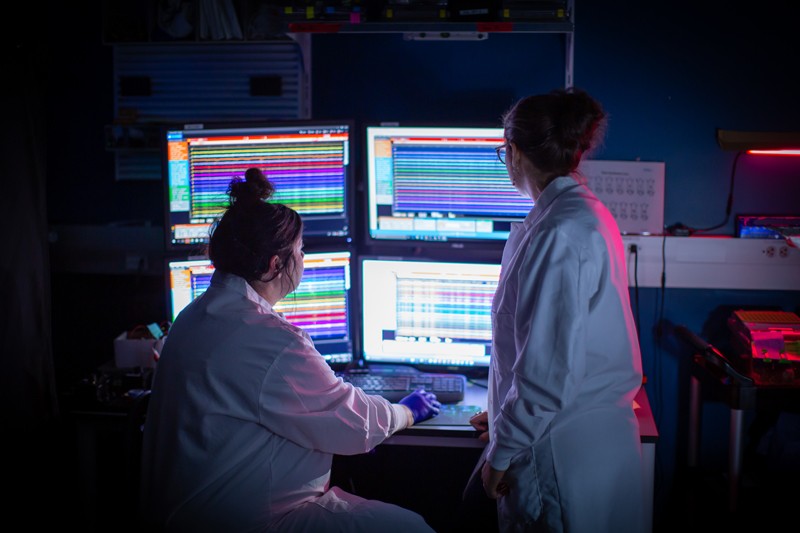 Two female researchers in white coats look at four computer monitors showing colourful data charts