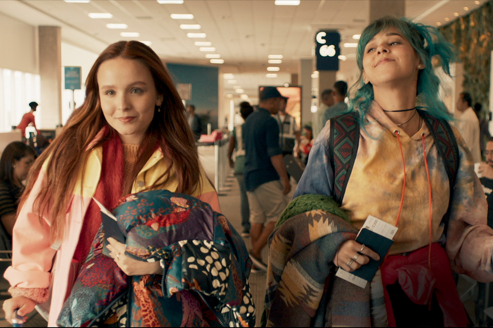 The image shows Larissa and Thati walking into an airport with their bags and happiness.